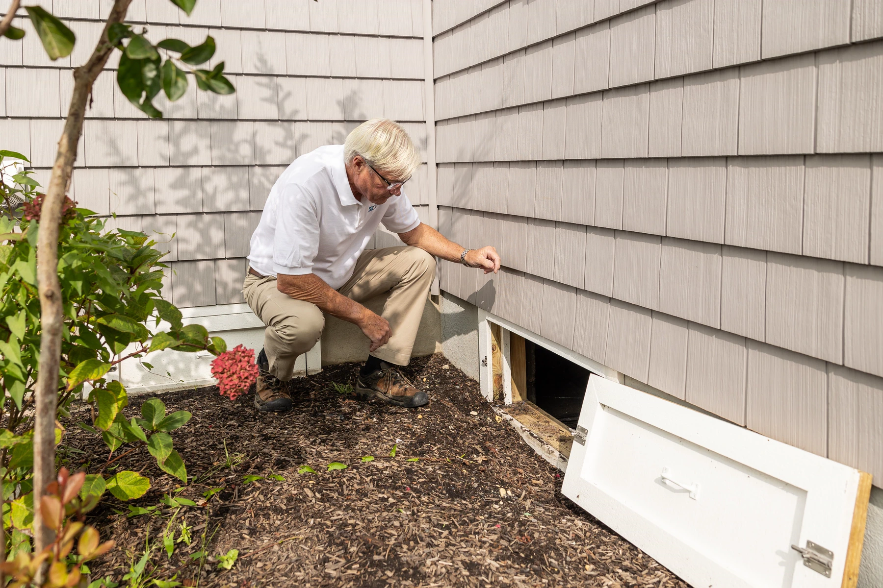 A resident inspects the crawlspace and foundation edge of a home